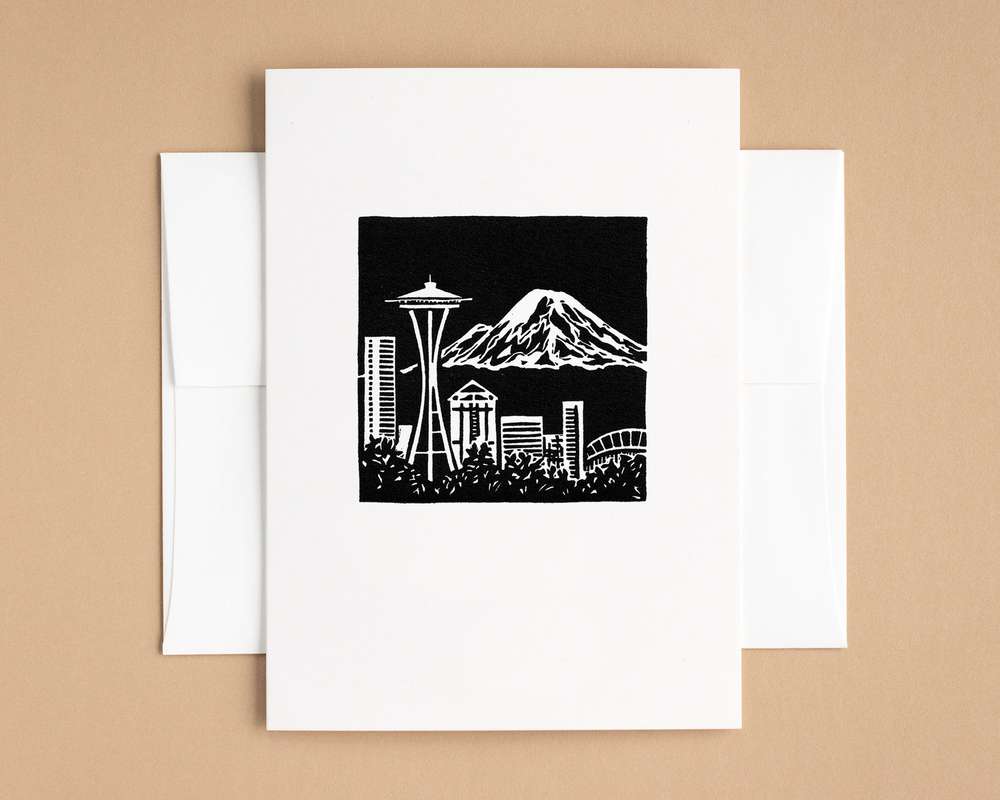 A vertical white card with a black and white depiction of the Seattle skyline and Mt Rainier rising in the distance. The card sits on top of a white envelope, which lies on top of a brown backdrop.